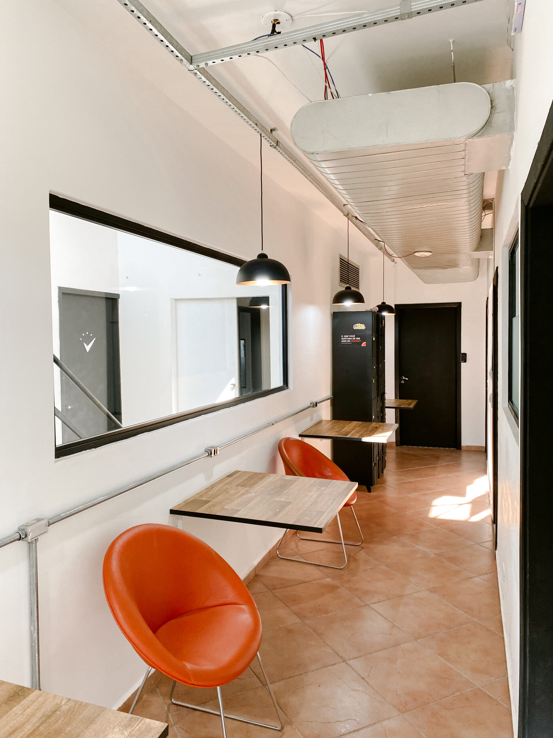 Corredor claro no Orange Coworking com mesas suspensas de madeira, poltronas laranjas, luminárias pretas e dutos aparentes no teto industrial.