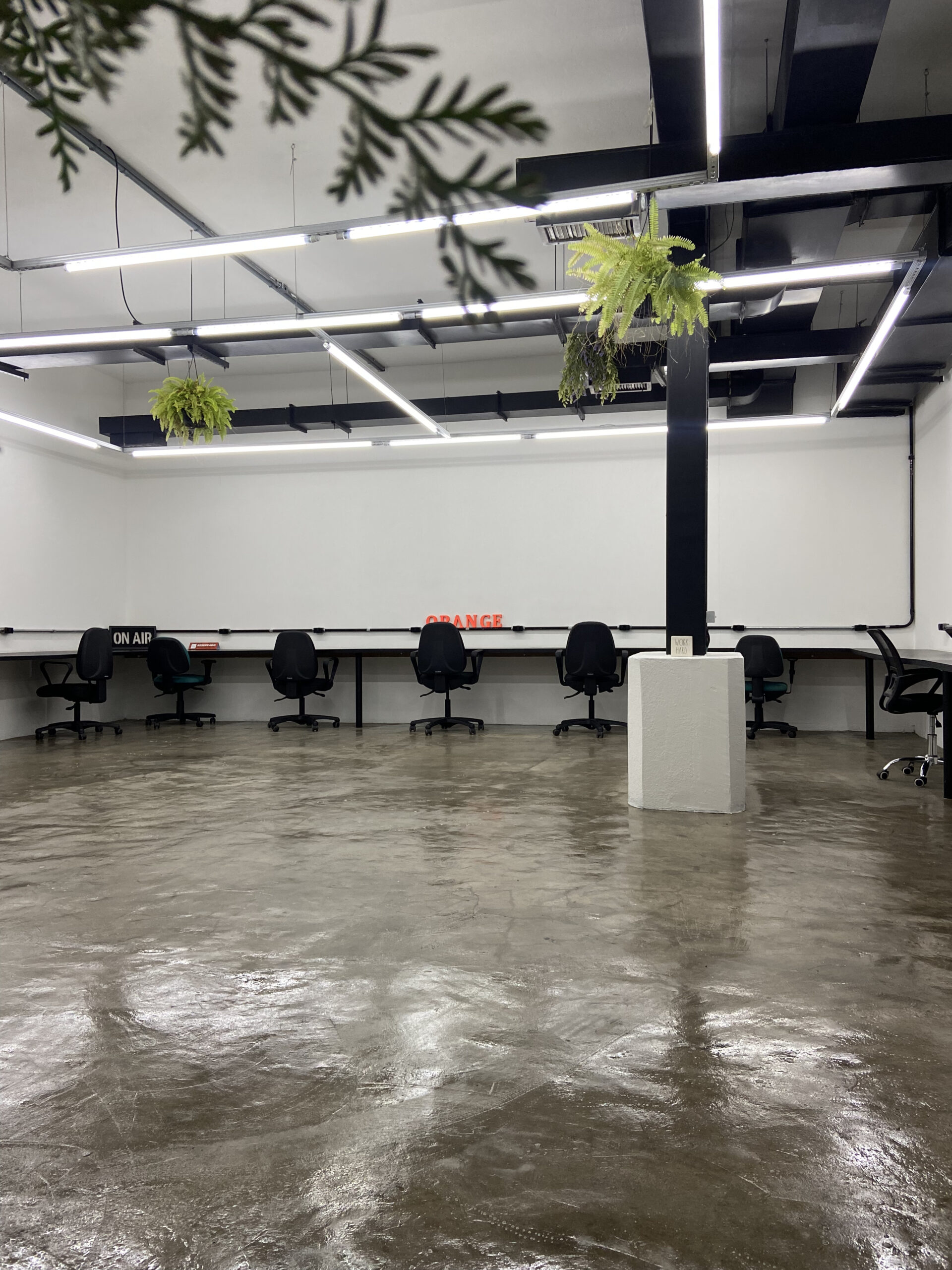 Amplo espaço de trabalho do Orange Coworking vazio com piso de cimento queimado brilhante, longa bancada branca, cadeiras pretas e teto industrial.