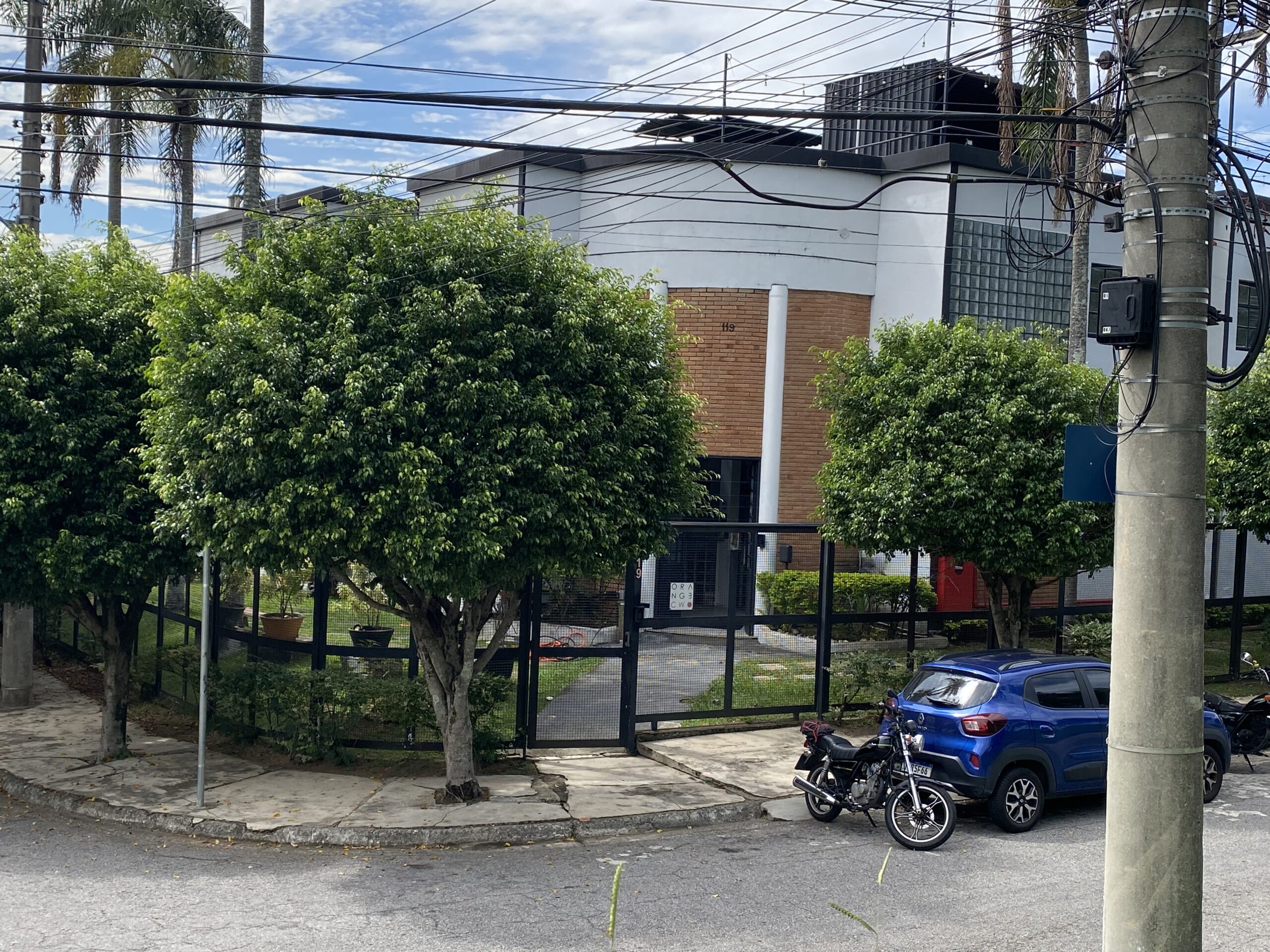 Vista da rua do Orange Coworking mostrando a calçada, grandes árvores verdes, grade preta e a fachada curva de tijolos com veículos parados.