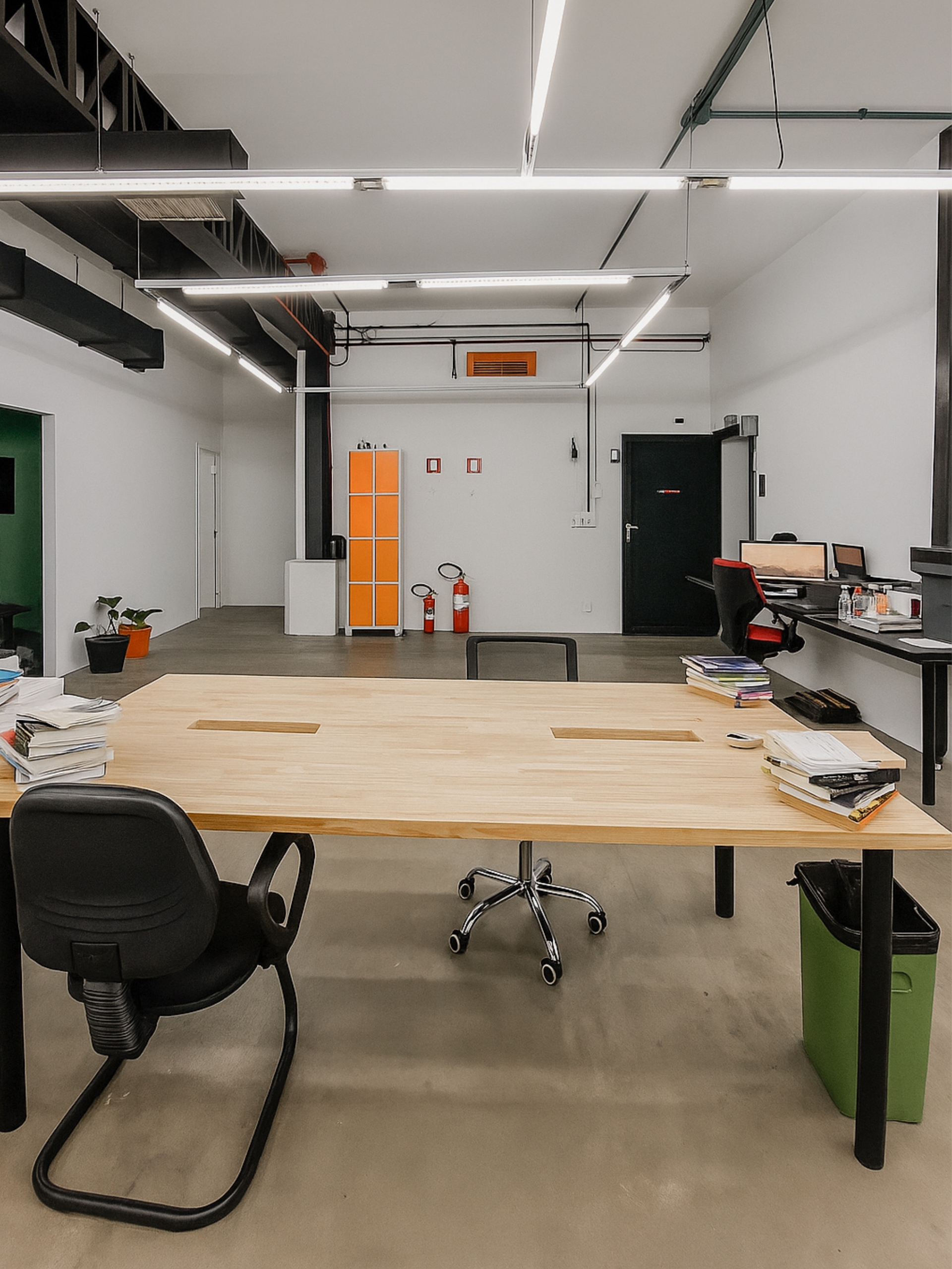 Espaço amplo do Orange Coworking com estilo industrial, teto aparente, iluminação linear, mesa de madeira em destaque e armário laranja.