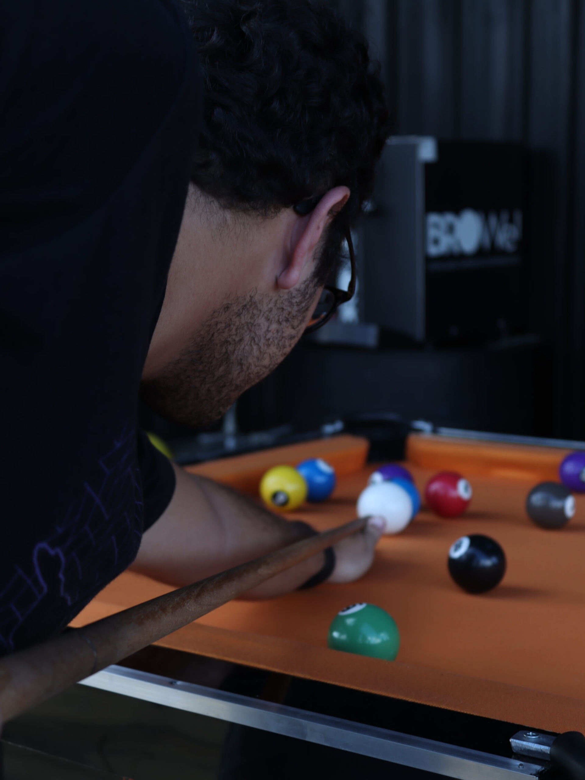 Orange Coworking Espaço para Eventos 1 Close-up de um homem de barba e óculos jogando bilhar em uma mesa de sinuca com pano laranja na área de lazer do Orange Coworking. Bolas coloridas estão na mesa.
