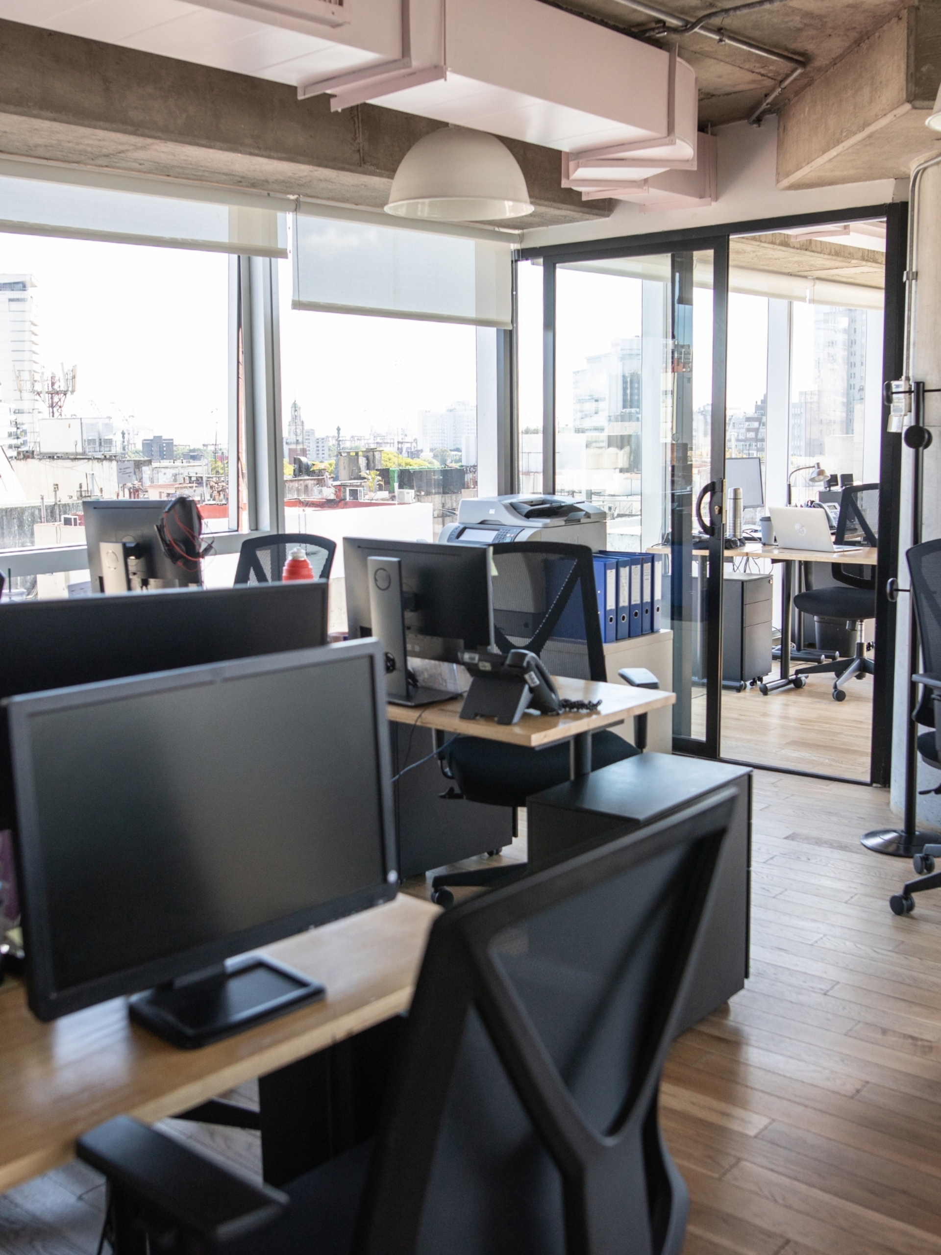 Orange Coworking Built to Suit 1 Escritório compartilhado do Orange Coworking com foco no estilo industrial, piso de madeira, monitores nas mesas e sala de vidro ao fundo.