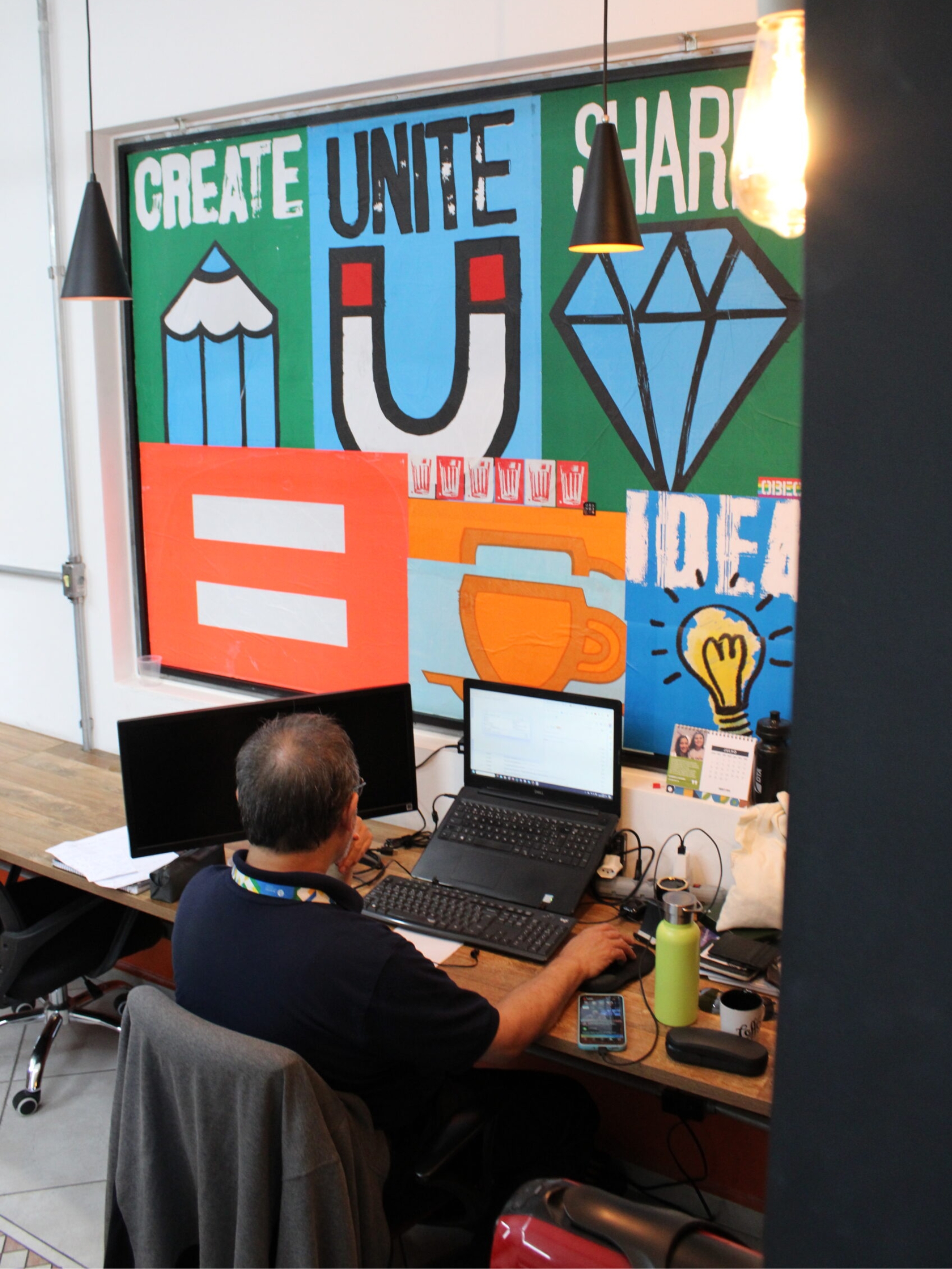 Orange Coworking Sala Privativa 1 Homem trabalhando em laptop no Orange Coworking. Ao fundo, painel colorido inspirador com as palavras Create, Unite, Share e ícones criativos.
