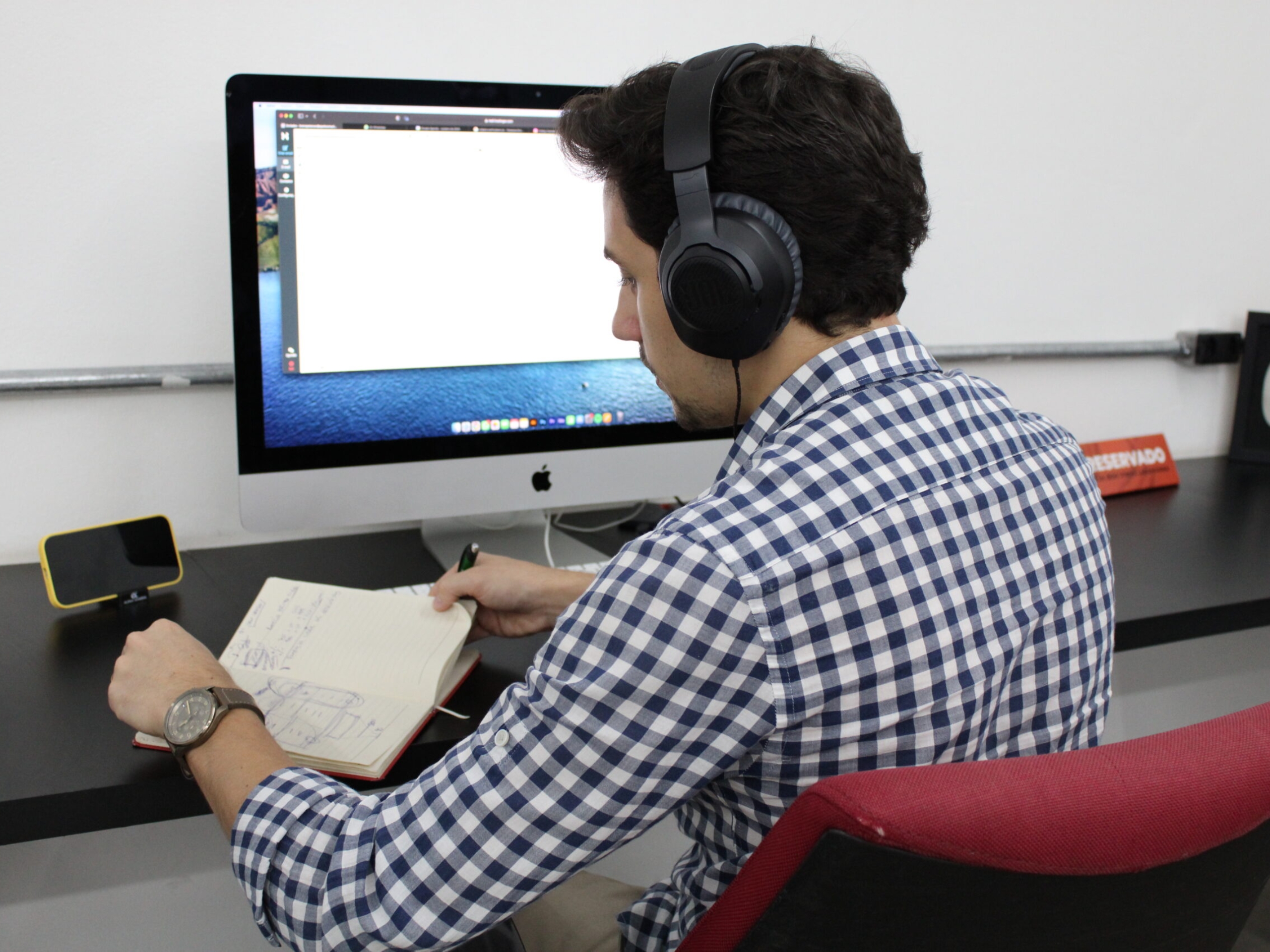 Homem de camisa xadrez e fones trabalhando focado no computador e fazendo anotações em uma mesa reservada no ambiente do Orange Coworking.