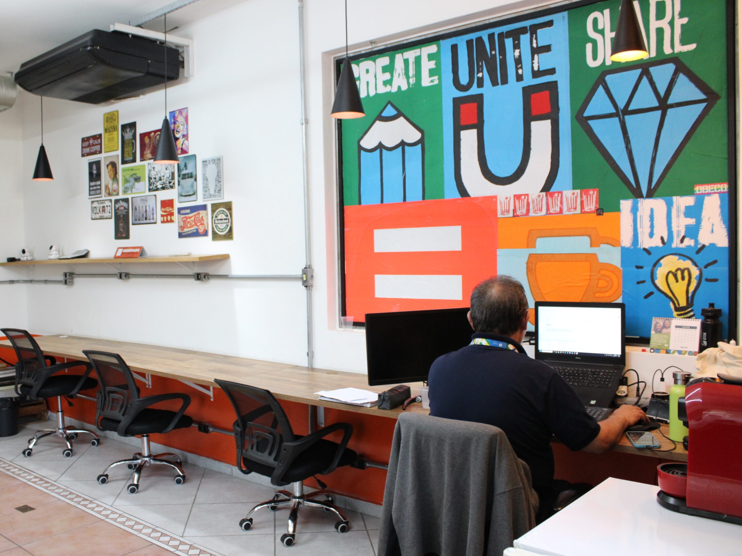 Orange Coworking Sala Privativa 4 Ampla estação de trabalho no Orange Coworking com longa mesa de madeira, cadeiras pretas, mural colorido inspirador e homem usando notebook.