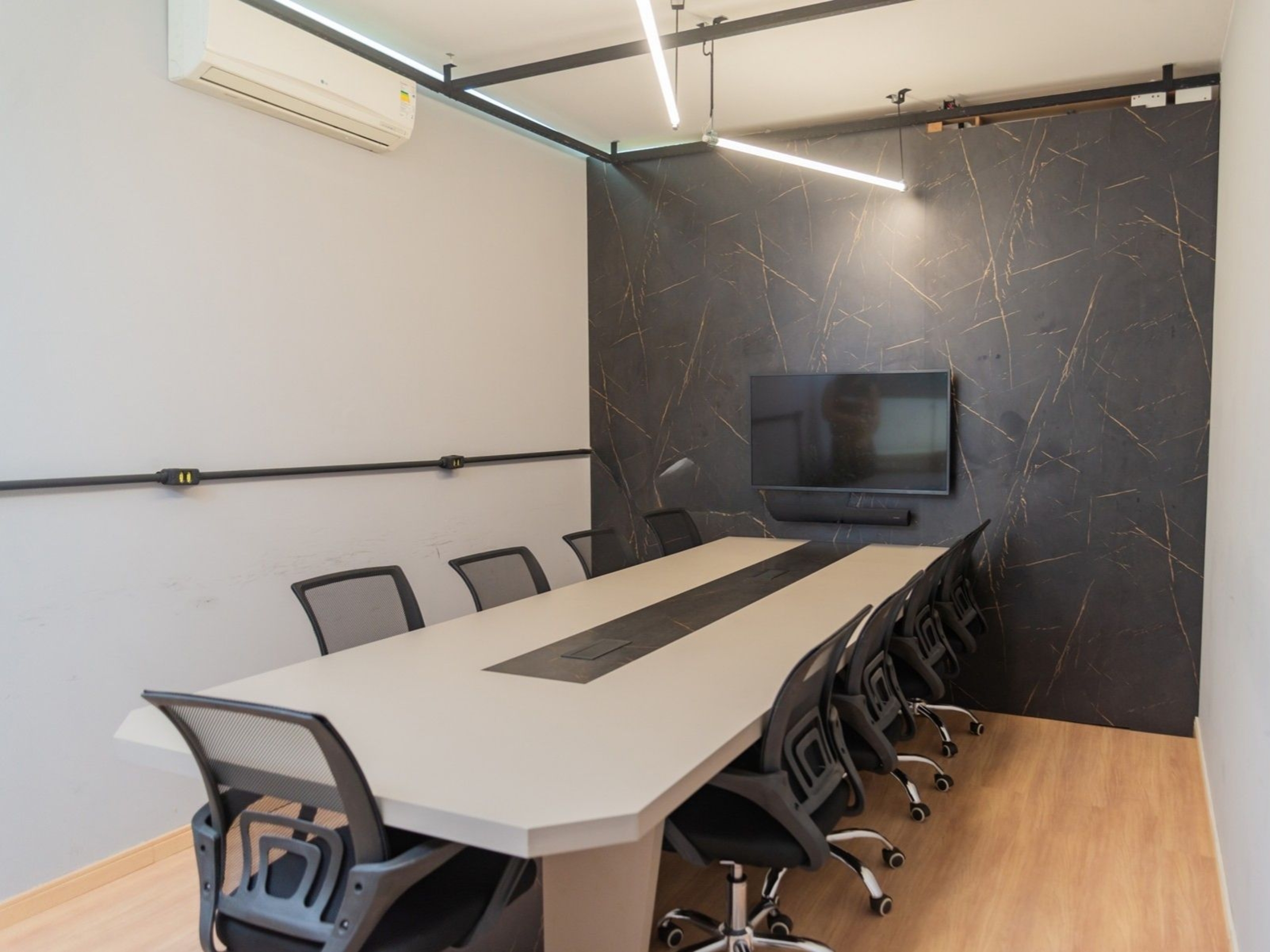 Elegante sala de reuniões no Orange Coworking destacando mesa clara, cadeiras giratórias, piso de madeira e TV instalada em painel escuro.