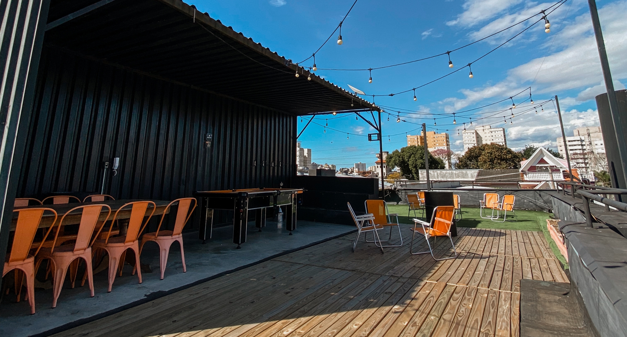 Terraço ao ar livre do Orange Coworking em dia ensolarado, decorado com deck de madeira, grama sintética, cadeiras laranjas de lazer, mesa de pebolim e varal de luzes.